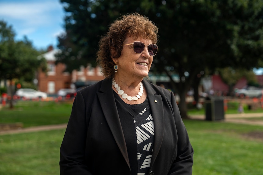 A woman wearing black smiles in a park with dark sunglasses and a white necklace.