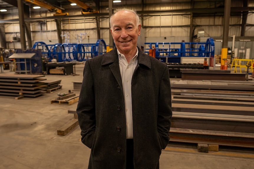 A man wearing a jacket and collared shirt stands in a factory.