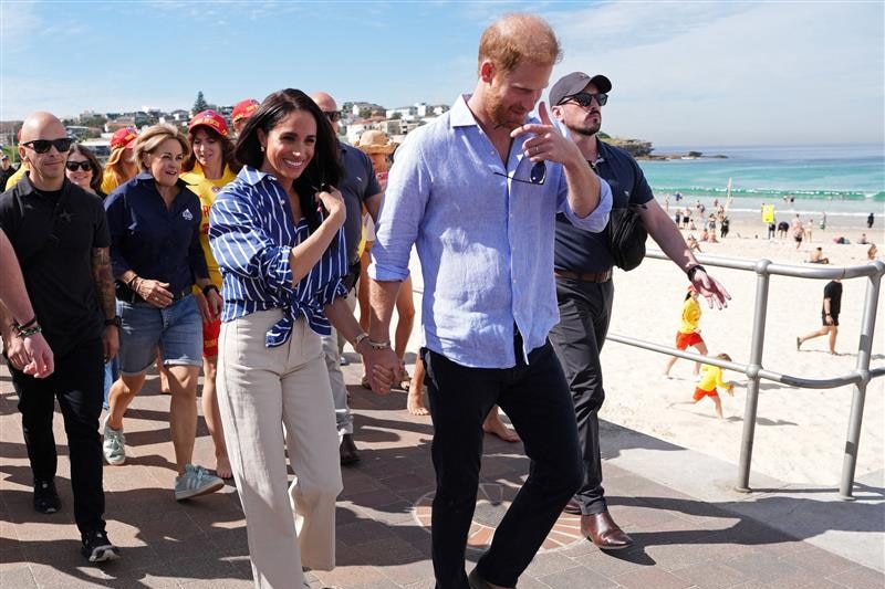 Harry and Meghan walk along Bondi Beach with surf life savers behind them.