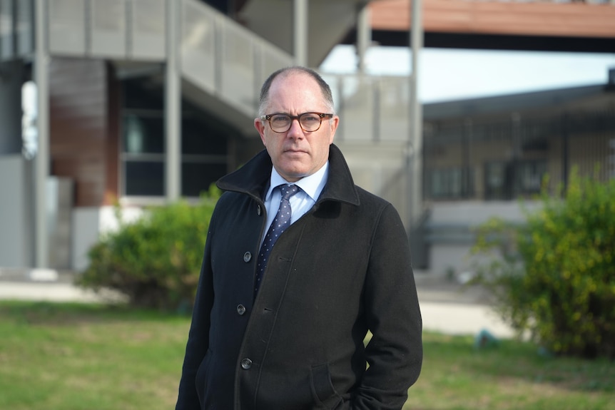A man with black and grey hair wearing glasses, a blue shirt and tie stands outside with his hand in the pocket of a black coat.
