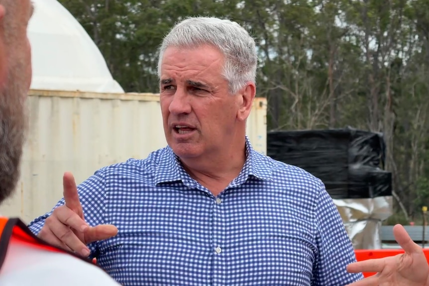 A grey-haired man gestures as he speaks at an industrial site.