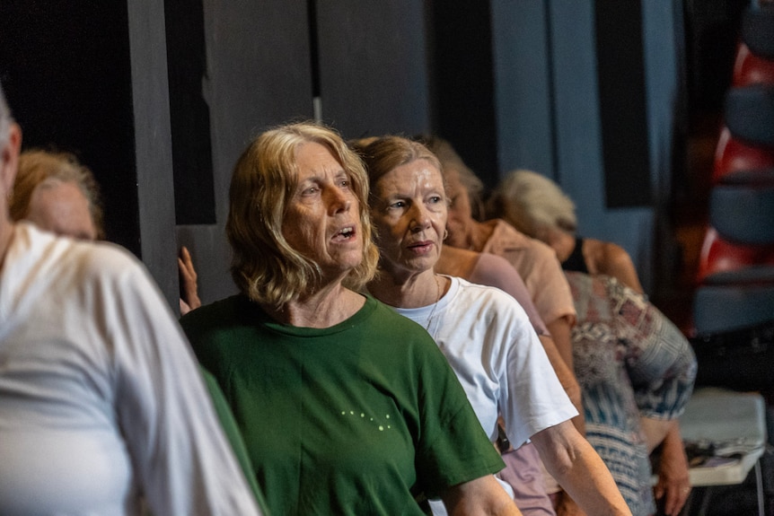 A line of six women lean with one hand up against a wall practicing a dance.