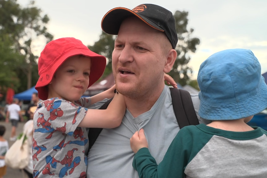 A man in a cap carries two children in his arms. Behind him a small crowd gathers looking away from camera
