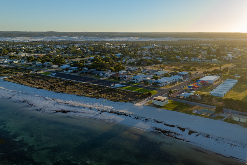 An aerial view shows the beach in the left corner and street sand buildings. Sand dunes and coastal scrub in the background.
