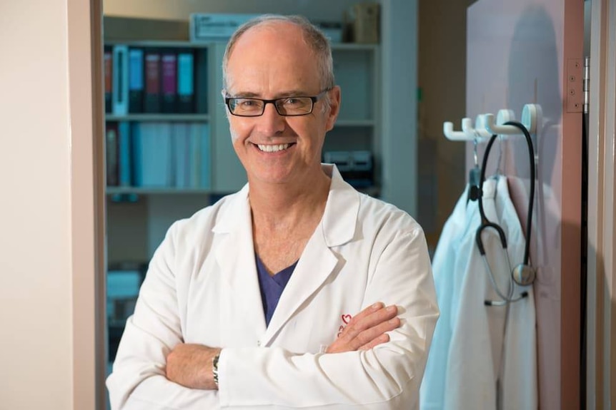 A man in a white lab coat over scrubs smiles at the camera.