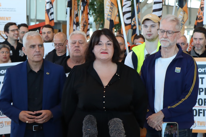 a woman in a black dress standing in front of a crowd of union members speaking to microphones