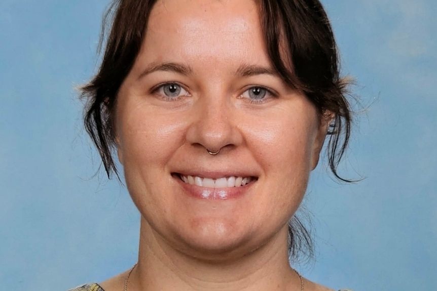 Woman with brown bangs smiling at camera in professional teacher photo, blue background.