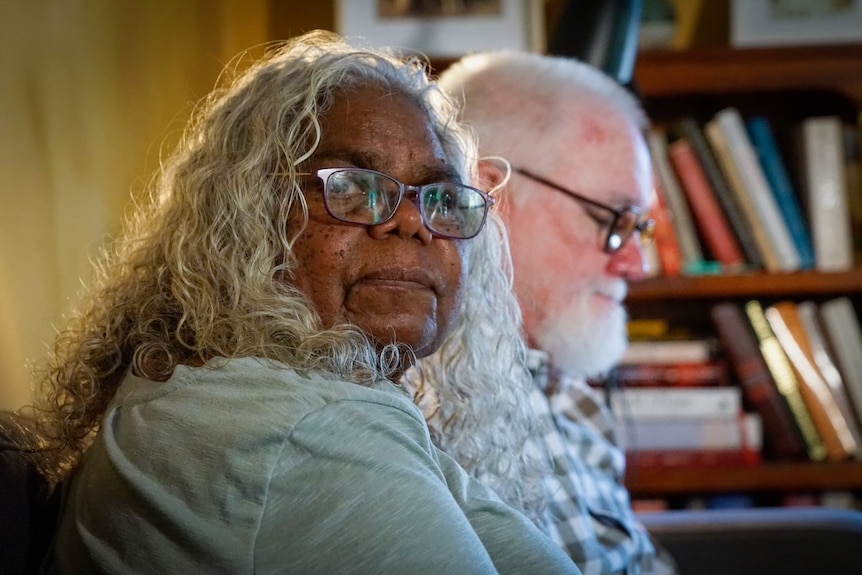 A woman looks seriously at the camera. She has curly grey, shoulder-length hair. A man is sitting next to her.