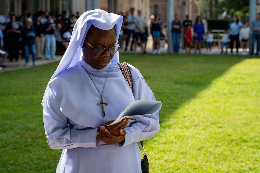 A woman dressed all in white with a cross stands on a lawn