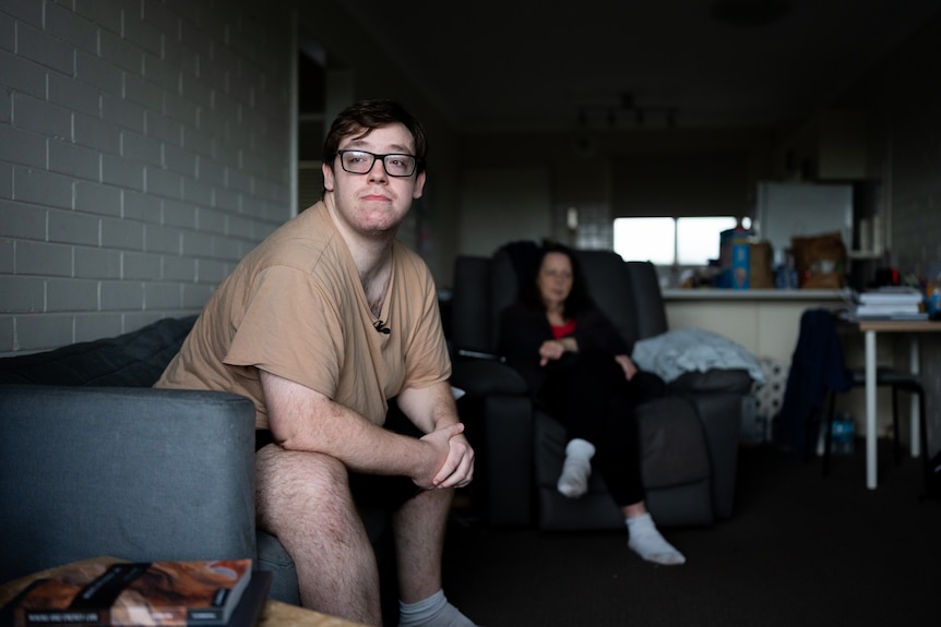 Brett and his mother sit on the couch in a dimly lit, small apartment living room.