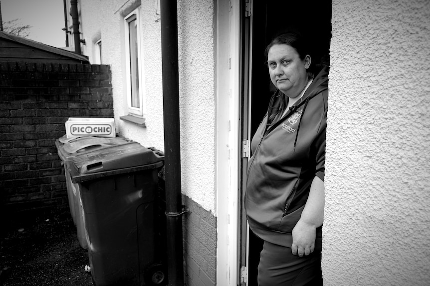 A woman standing in a doorway with bins outside leaning against the wall.
