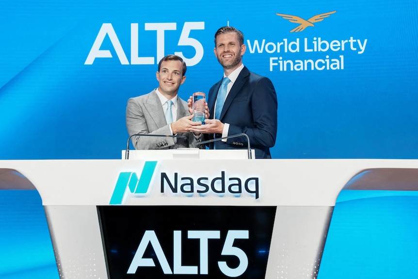 two white men in suits hold a trophy while standing behind a lectern that says nasdaq on it