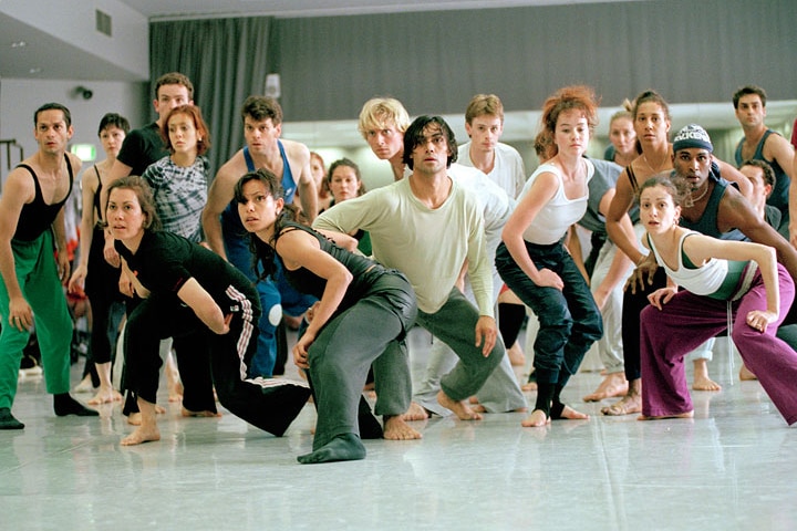 Frances Rings, an Indigenous dancer in her 30s, crouches, alert, in front of a group of other readied dancers.