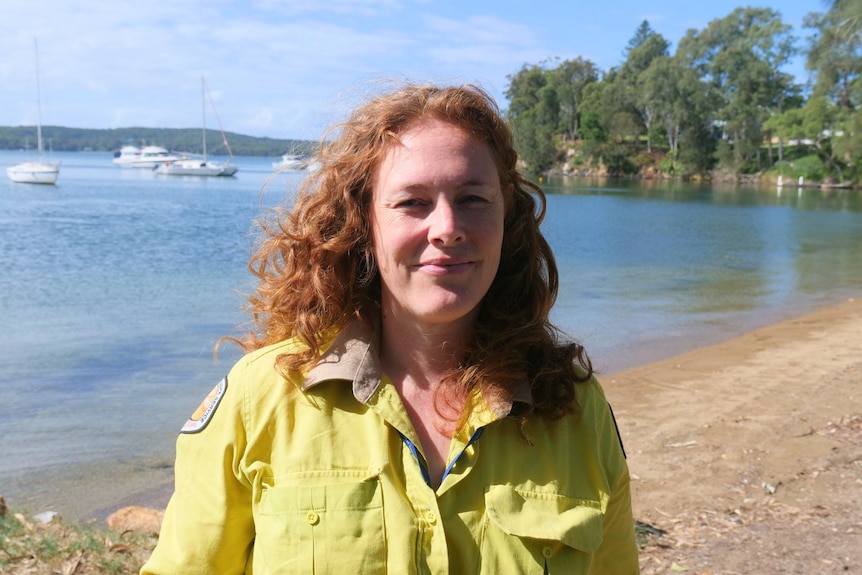 Nicola Booth, who has long curly hair, wears a yellow collared shirt. A lake is in the background.