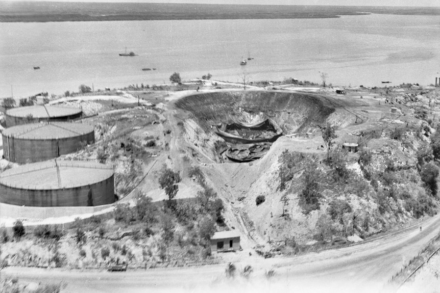 A black and white photo shows a huge crater in the ground, alongside three undamaged oil tanks located near the coastline.