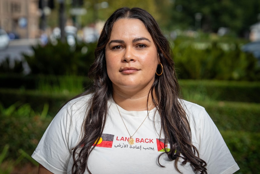 A woman with long brown hair wearing a white t-shirt with the words "LAND BACK" and an Aboriginal flag and a Palestinian flag.
