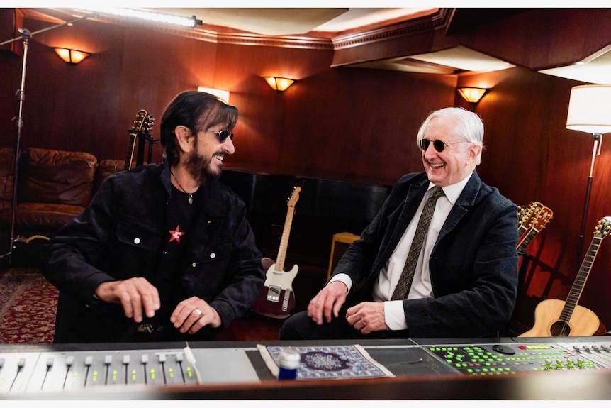 Ringo Starr and T Bone Burnett sit at a mixing desk in a wood panelled recording studio. They are both laughing.