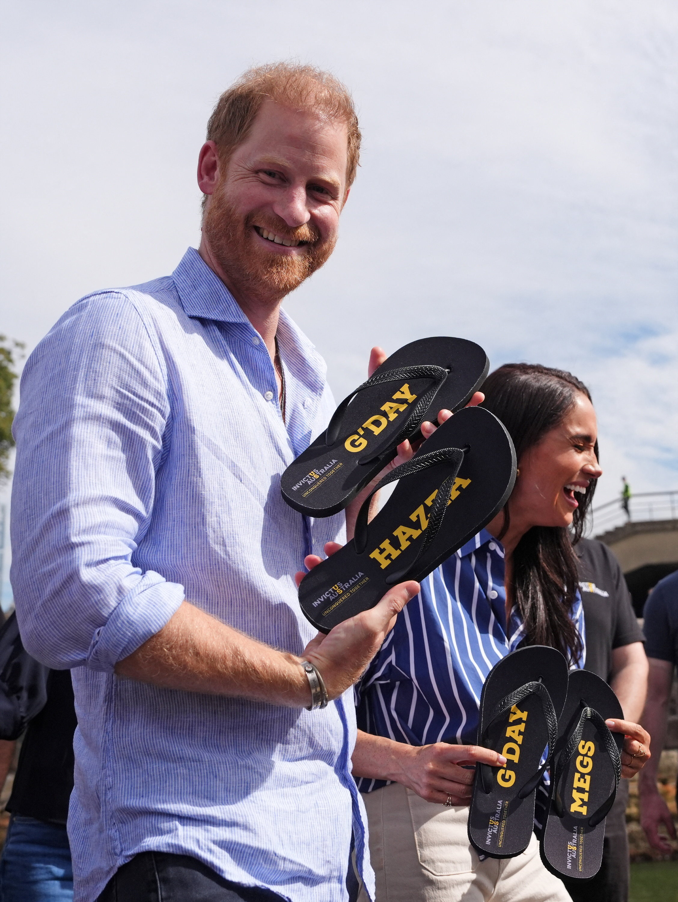 Prince Harry and Meghan laugh and hold black thongs that say 'G'day Hazza' and 'G'day Megs'