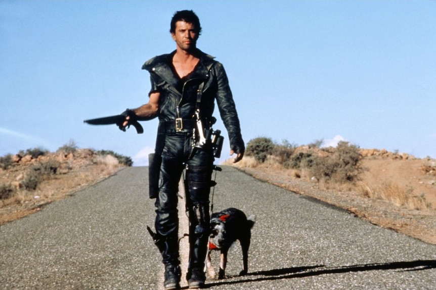 Man in leather walks down a road holding a sawnoff shot gun with dog next to him.