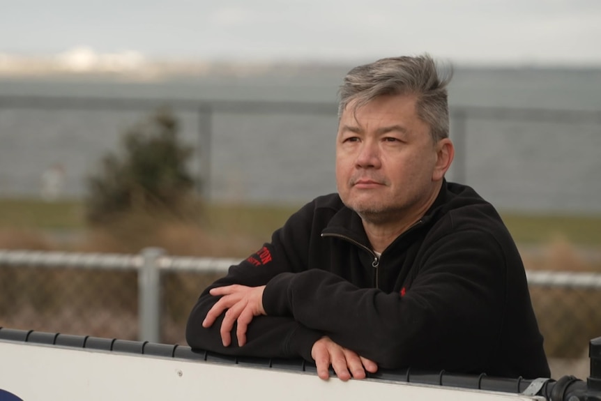 A man with his arms resting on a fence looks into the distance.