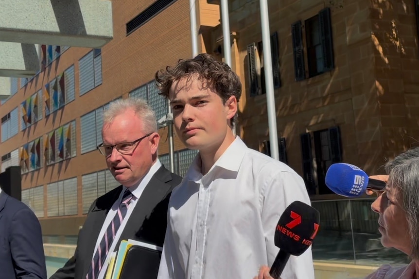 A man in a suit walks next to a younger man wearing a white shirt, while journalists nearby hold microphones