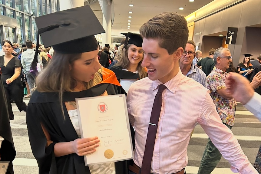 A young lady and in a graduation hat next to a young man in a business shirt and tie in a foyer at a university.
