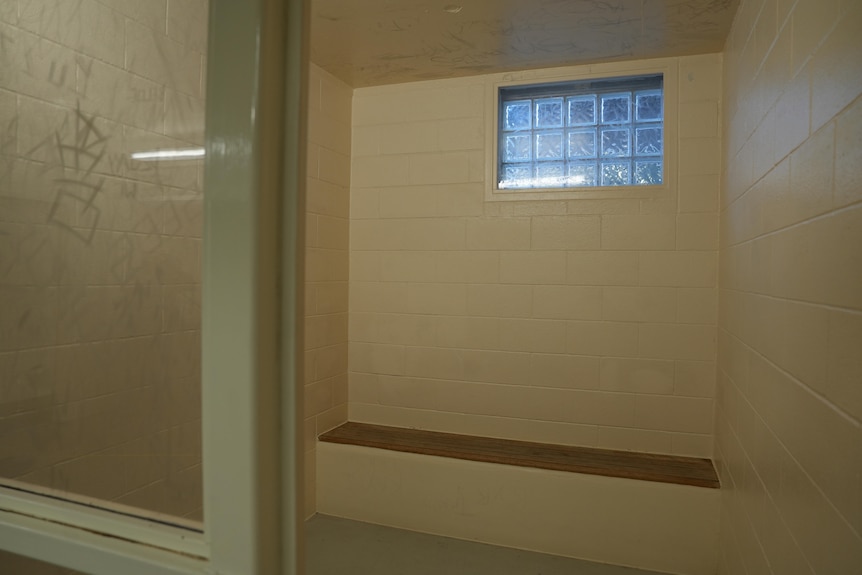 A small cell with a bench under an enclosed glass window. A Persplex wall next to the opened door