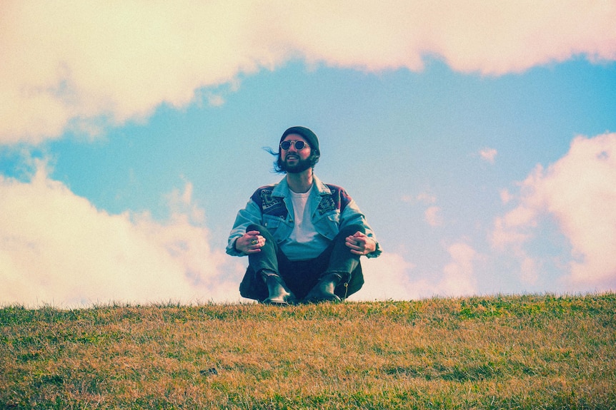 Man in a beanie and denim jacket sits on a crass hill and looks off into the distance. The clouds are edited pink tone. 