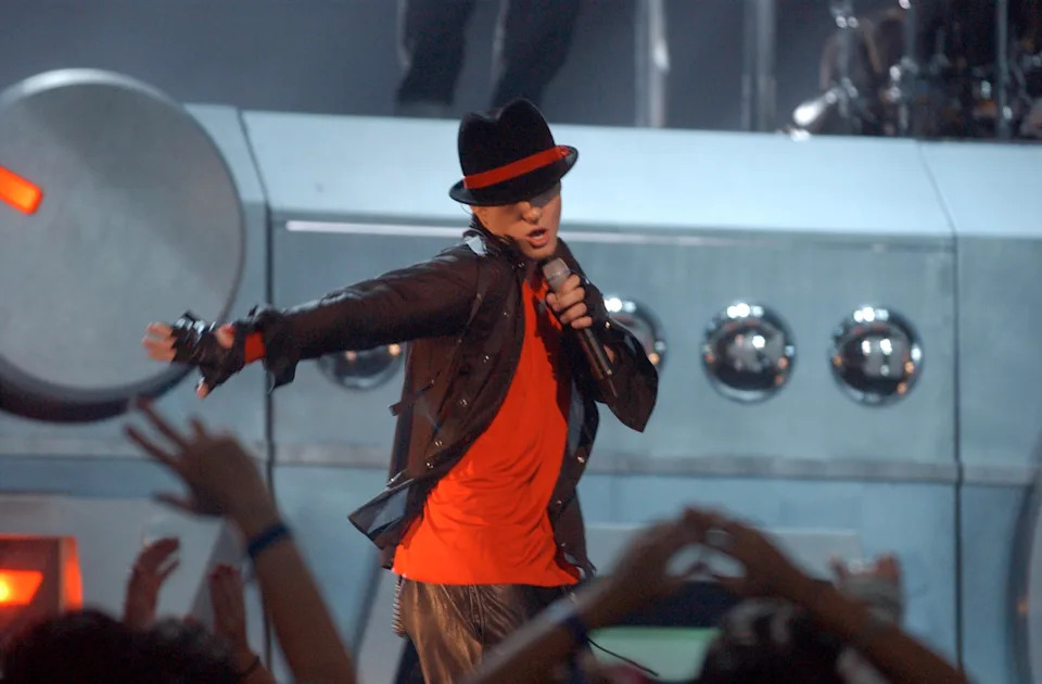 Performer on stage in a dynamic pose, wearing a stylish jacket, fedora, and sunglasses, holding a microphone during a concert