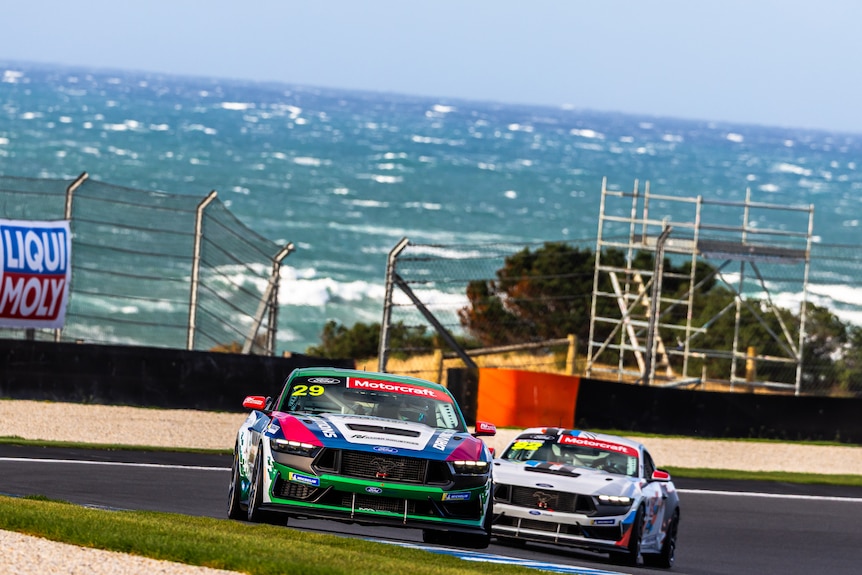 Two race cars come around a bend with a choppy ocean behind them.