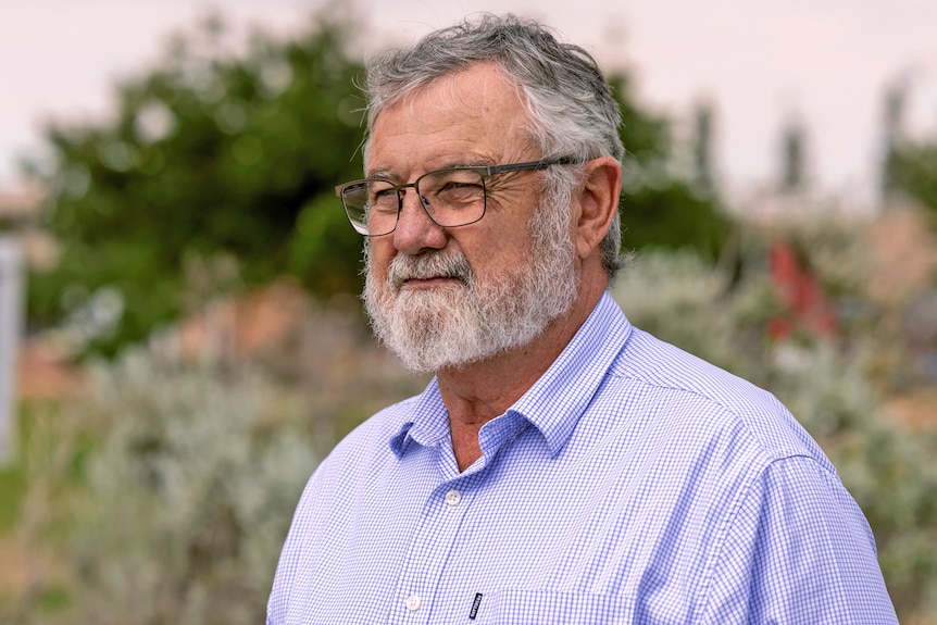 An older, bearded man in glasses stands in front of some shrubs.