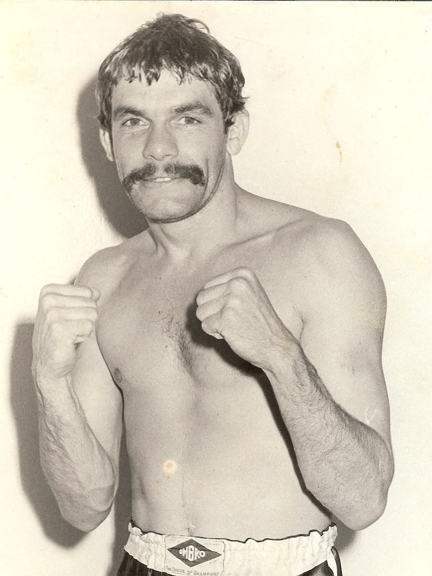 A black and white image of a man with short dark hair standing with his fists up, wearing boxing gloves.