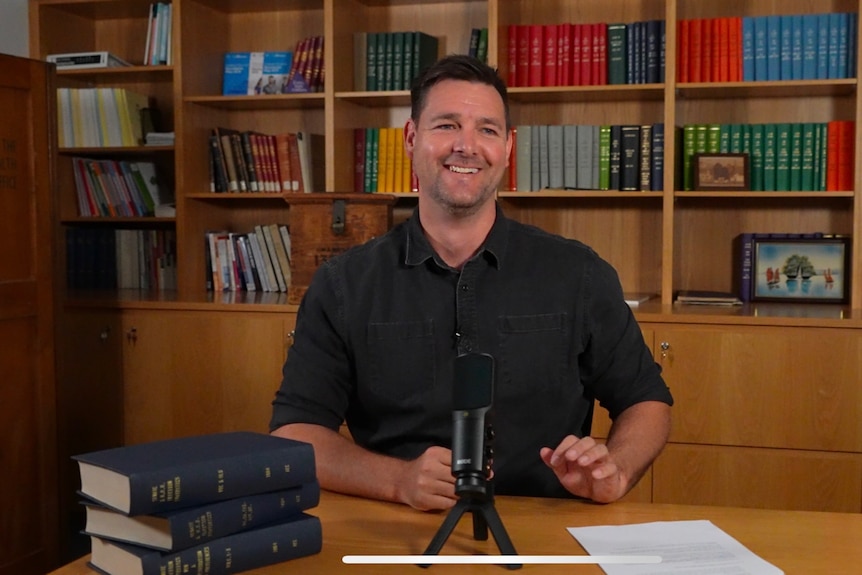A man with dark hair sits in front of a microphone against a backdrop of books.