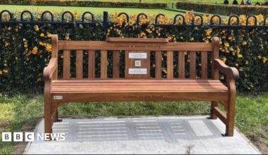 A bench placed on a concrete slab. It is placed in front of a metal fence.