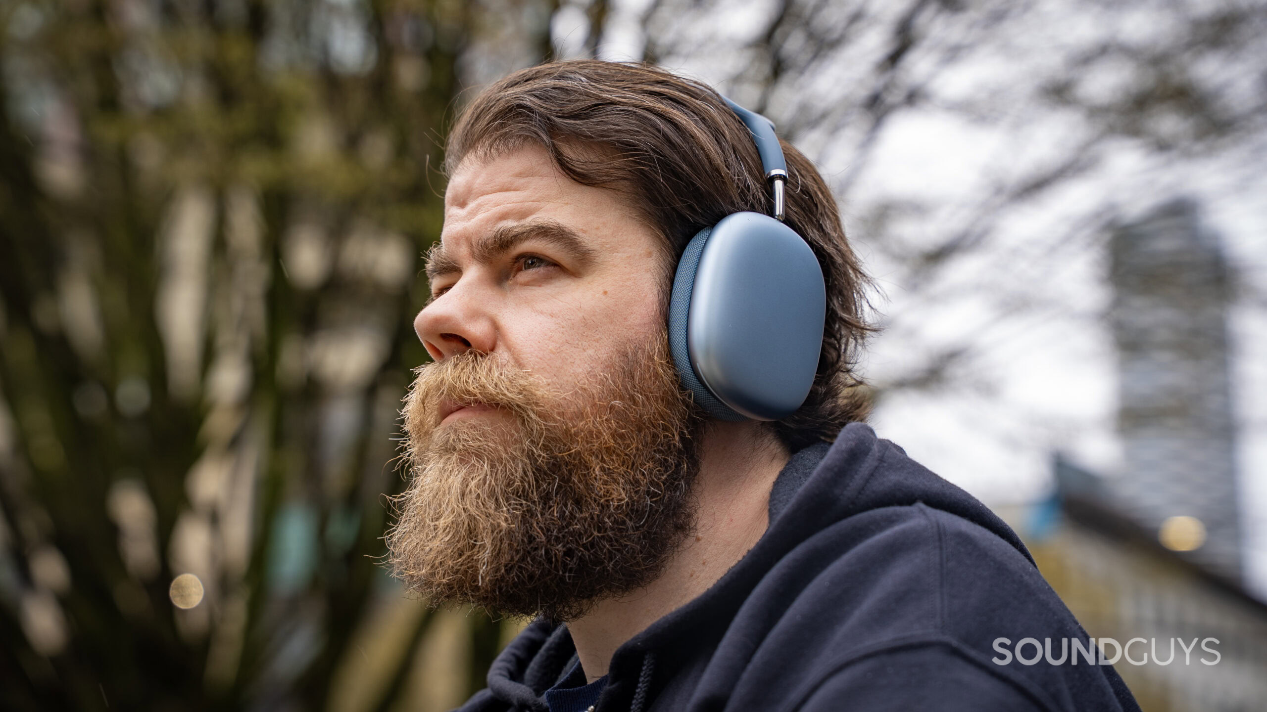 apple airpods max 2 close up man weawring in front of tree Close up of man wearing the Apple AirPods Max 2 headphones as he looks off into the distance.