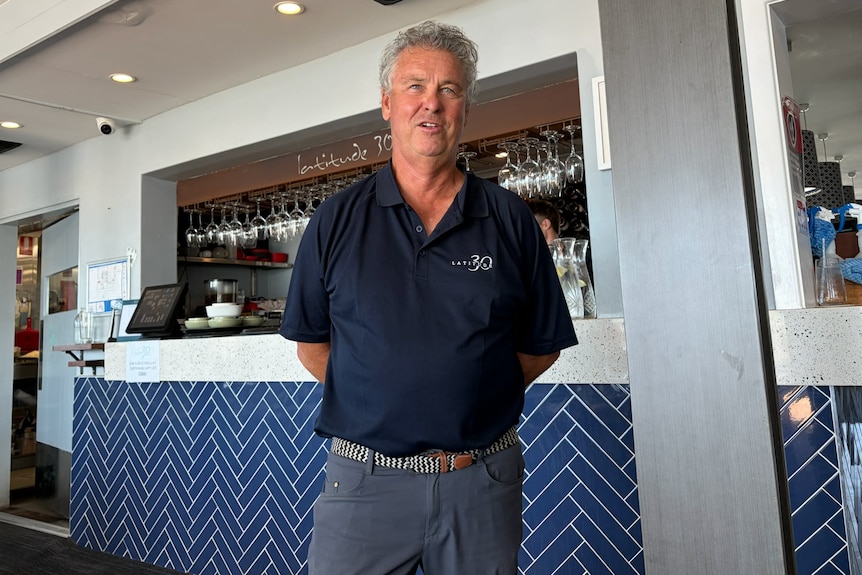 A middle aged man with silver hair and a blue t-shirt stands in front of a restaurant bar