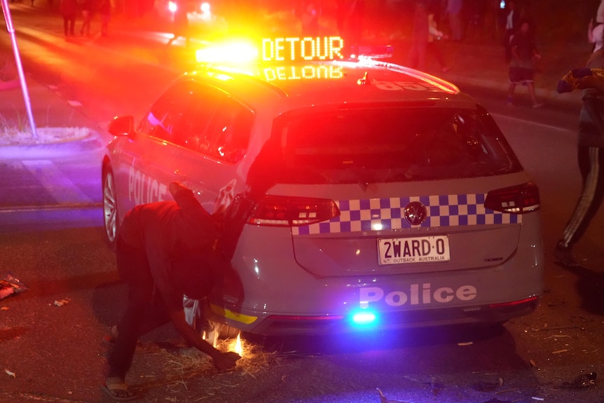 A man using a lighter to attempt to ignite dry grass under a police vehicle at night.
