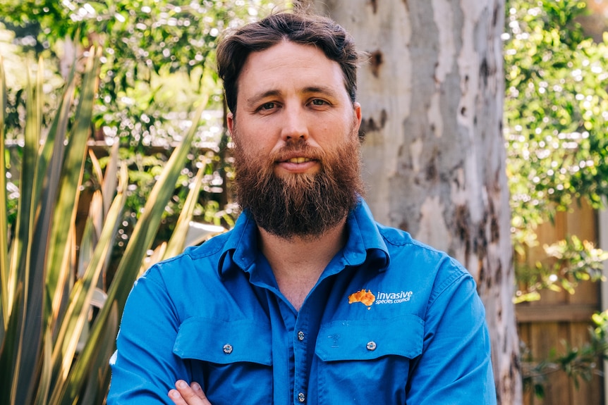 A bearded man with folded arms stands in front of the trunk of a large gum tree