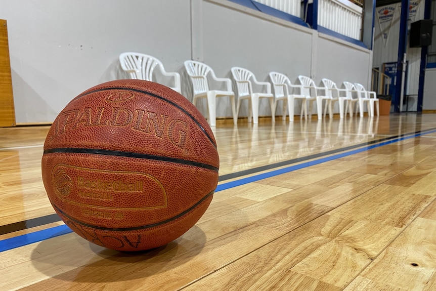 A basketball on an indoor basketball court placed in front of a row of empty bench chairs. 