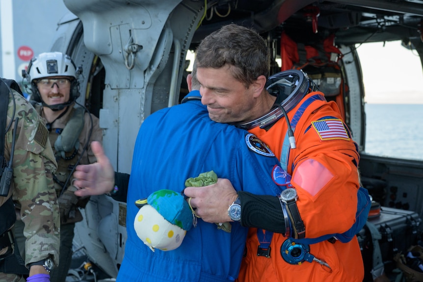 A man in a space suit smiles and hugs a man in blue overalls whose back is towards the camera