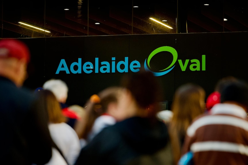 The backs of heads walking towards a lit up sign that says Adelaide Oval. 