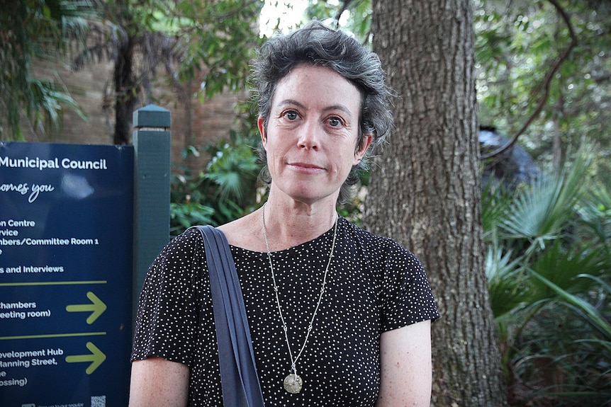 Woman with grey hair and dark clothing in front of council sign and trees.