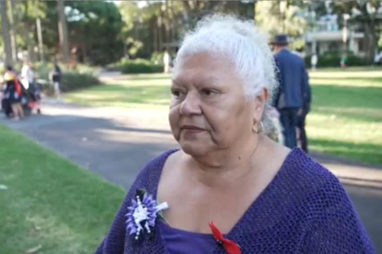 An Aboriginal woman with grey hair and a purple top with a broach on one side standing in a park