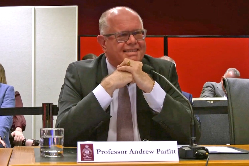 An older balding man sitting at a parliamentary evidence desk with a glass of water, microphone and name plate.