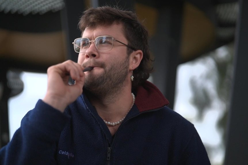 A man with short brown hair hits a vape outdoors.