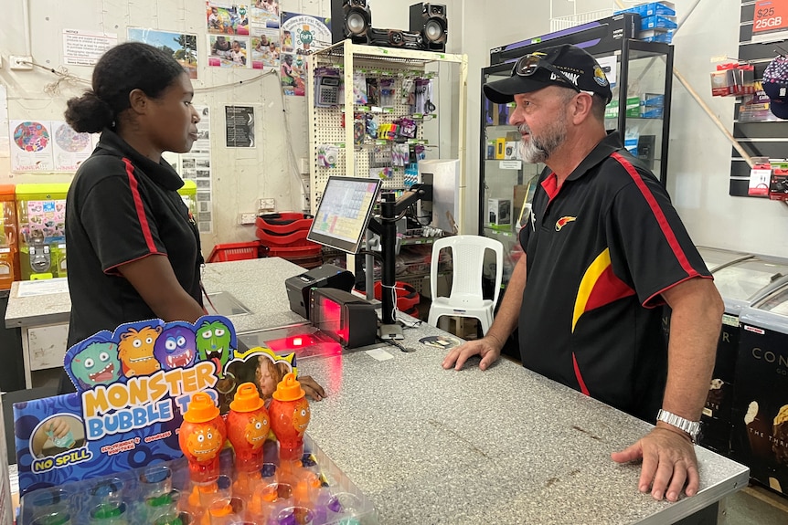 Alastair King stands at the checkout register of a small grocery store, speaking to the worker behind the counter.
