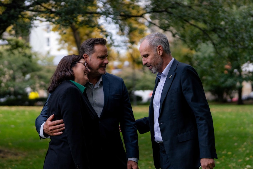 A woman and man in formal wear embrace, another man with grey hair and beard extends an arm.