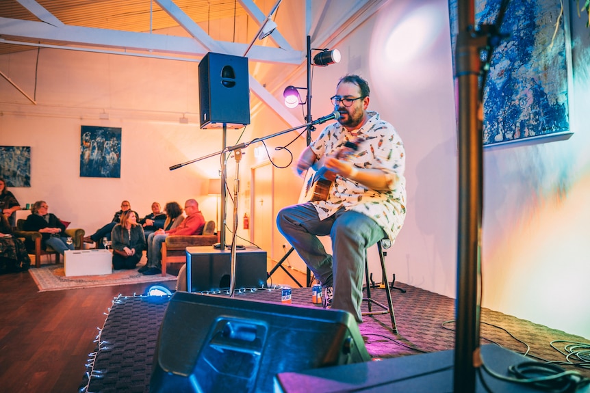 A man sitting on a seat with a microphone on a stage sings and plays the guitar.