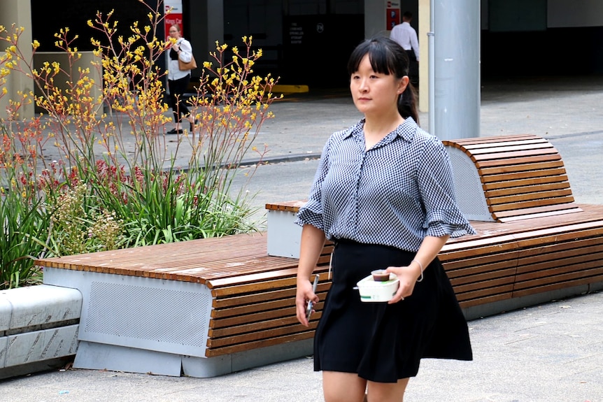 Mimi Yeung in a shirt and skirt, walking in front of a bench carrying her lunch.