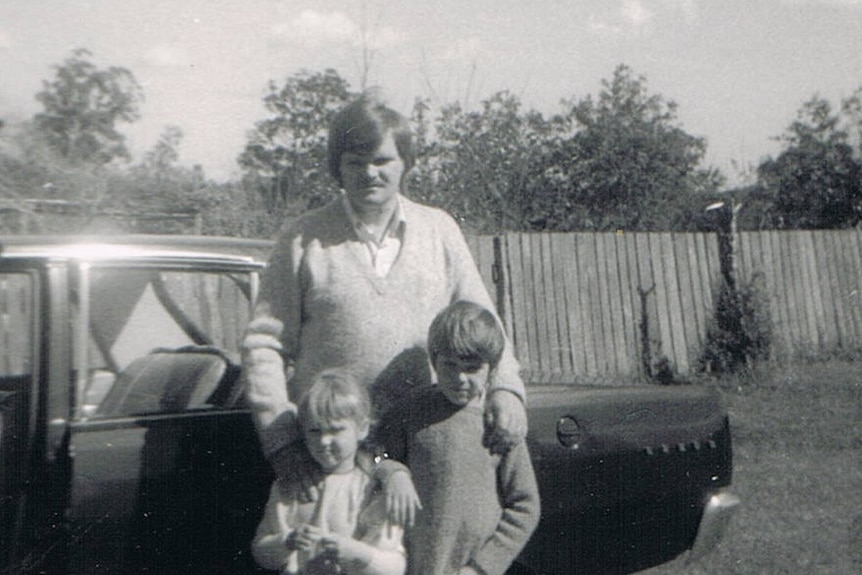 A black and white photo of a young man with two kids next to a car.